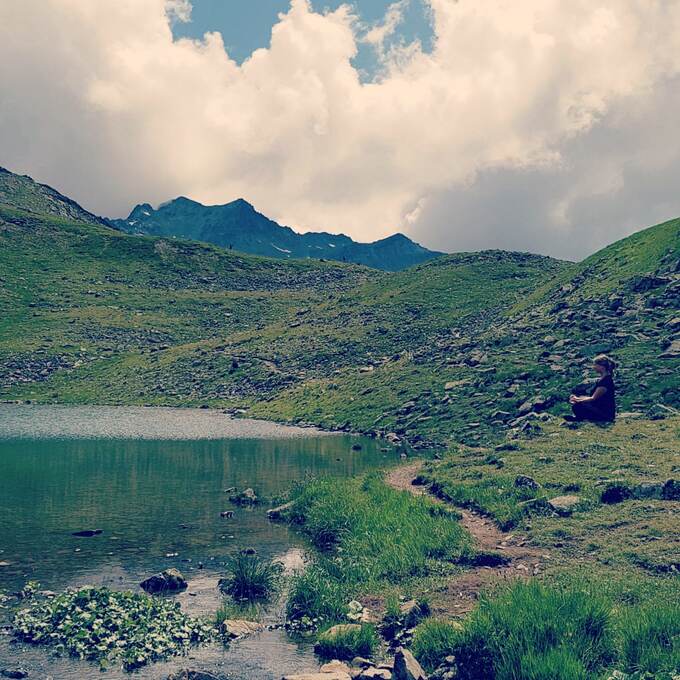 Person sitzt am Ufer eines Bergsees, umgeben von gr&uuml;nen Wiesen und Bergen unter einem bew&ouml;lkten Himmel.