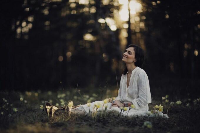 Frau im wei&szlig;en Kleid sitzt auf einer Wiese mit Grazien, umgeben von verschwommenen B&auml;umen im Hintergrund.