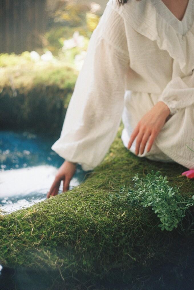 Hand einer Person in einem wei&szlig;en Kleid ber&uuml;hrt moosbedeckten Wall am Rand eines blauen Gew&auml;ssers.
