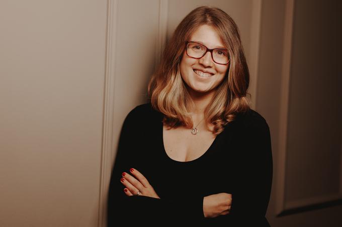 Portr&auml;t einer l&auml;chelnden Frau mit Brille, die ihre Arme vor einem grauen Hintergrund verschr&auml;nkt.