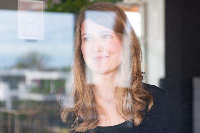 Portr&auml;t einer Frau mit langen Haaren, die durch ein Fenster l&auml;chelt, im Hintergrund leicht verschwommene Geb&auml;ude.